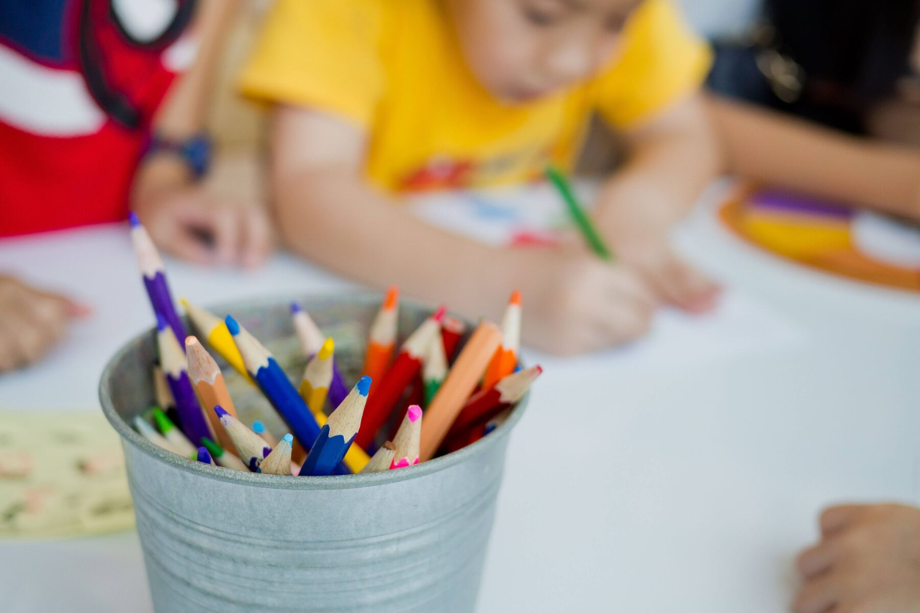 A Bucket of colored pencils sitting on a table, children coloring in the background