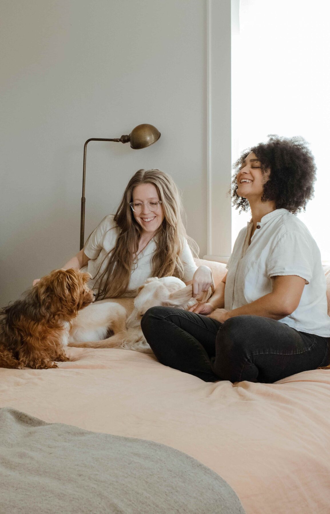 Two women sit on a bed smiling and petting two dogs. One woman has long hair and the other has curly hair. They appear relaxed and happy in a well-lit, cozy room with a lamp in the background.